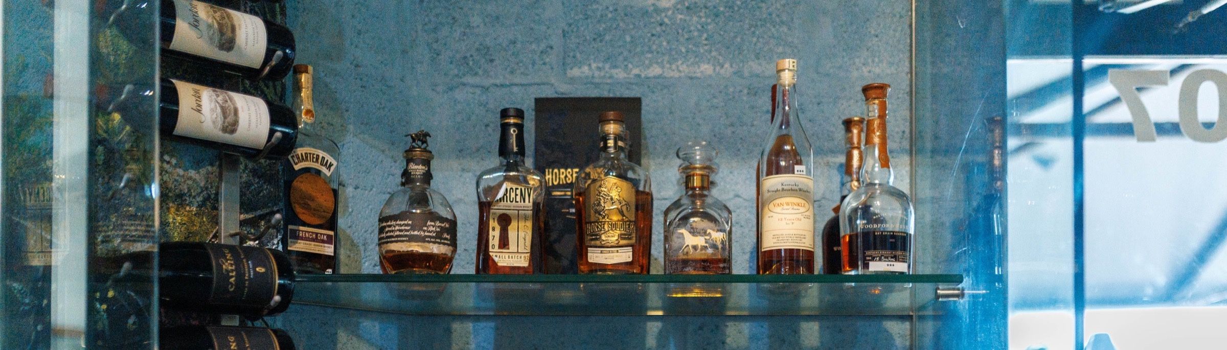 Glass shelves lined with bourbon, including Blanton's, Heaven's Door, High West and Knob Creek, beside a wine rack